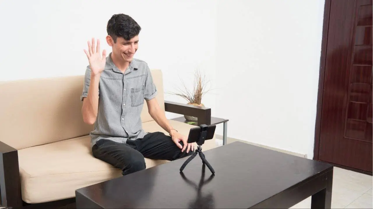A man sitting on a sofa, waving and smiling towards his phone placed on a tripod
